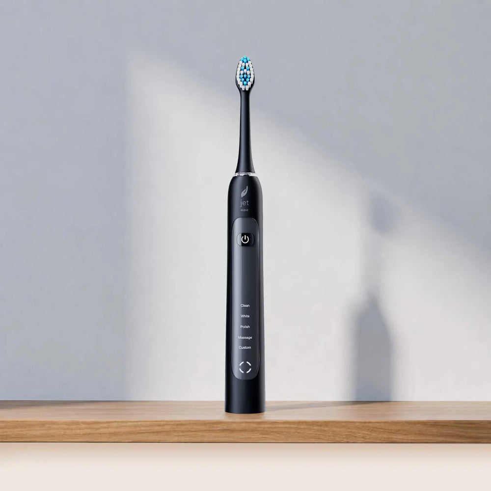 A sleek black electric toothbrush with blue and white bristles stands upright on a wooden surface against a light gray wall with soft shadows.