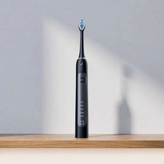 A sleek black electric toothbrush with blue and white bristles stands upright on a wooden surface against a light gray wall with soft shadows.